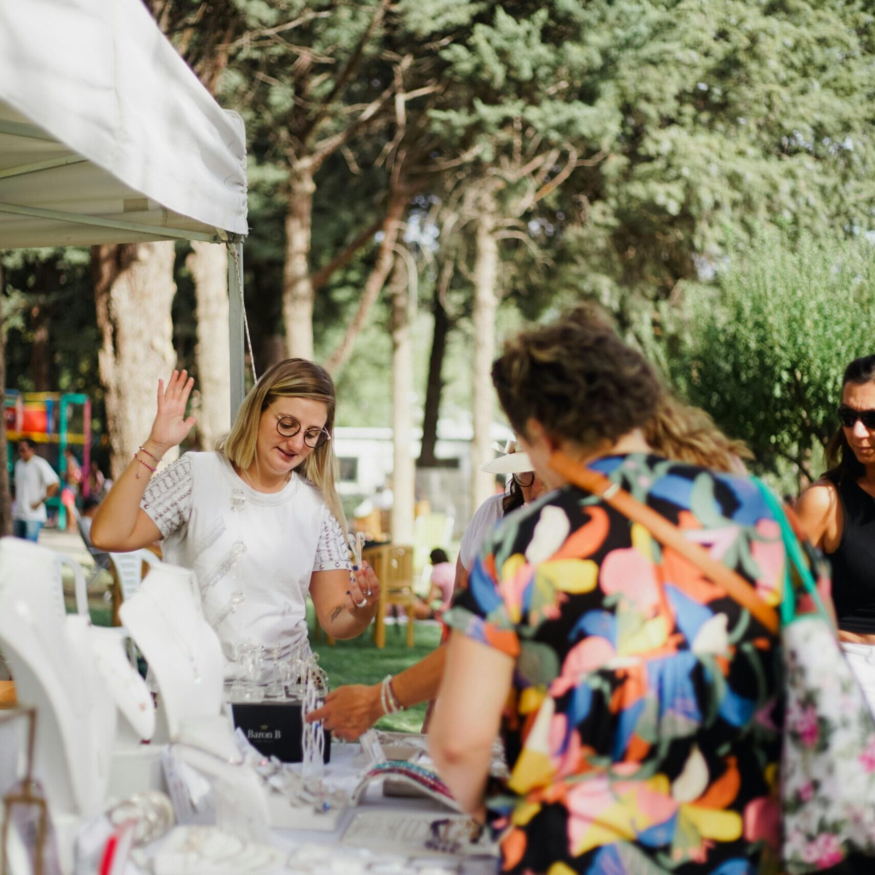 A lively scene of people shopping for jewelry at an outdoor market in a park during summer.