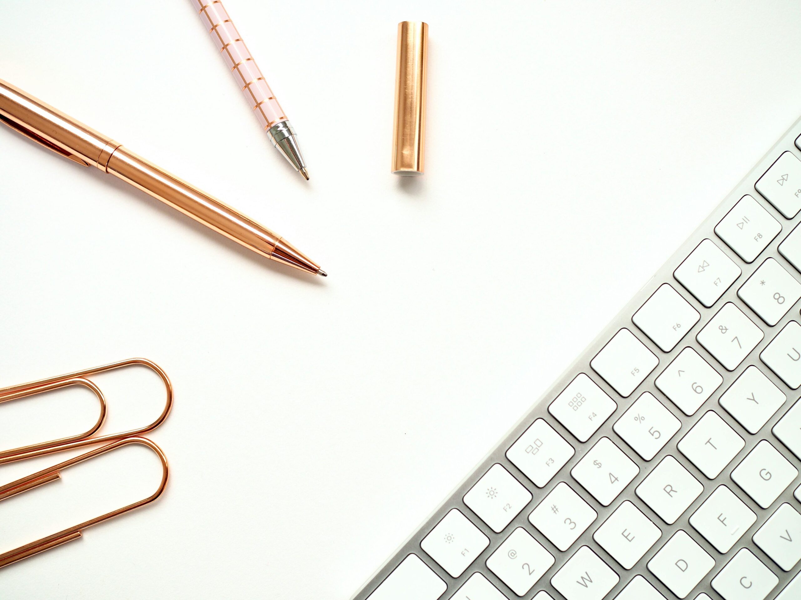 Elegant minimalist workspace featuring rose gold pens, paper clips, and a sleek keyboard for a stylish office setup.